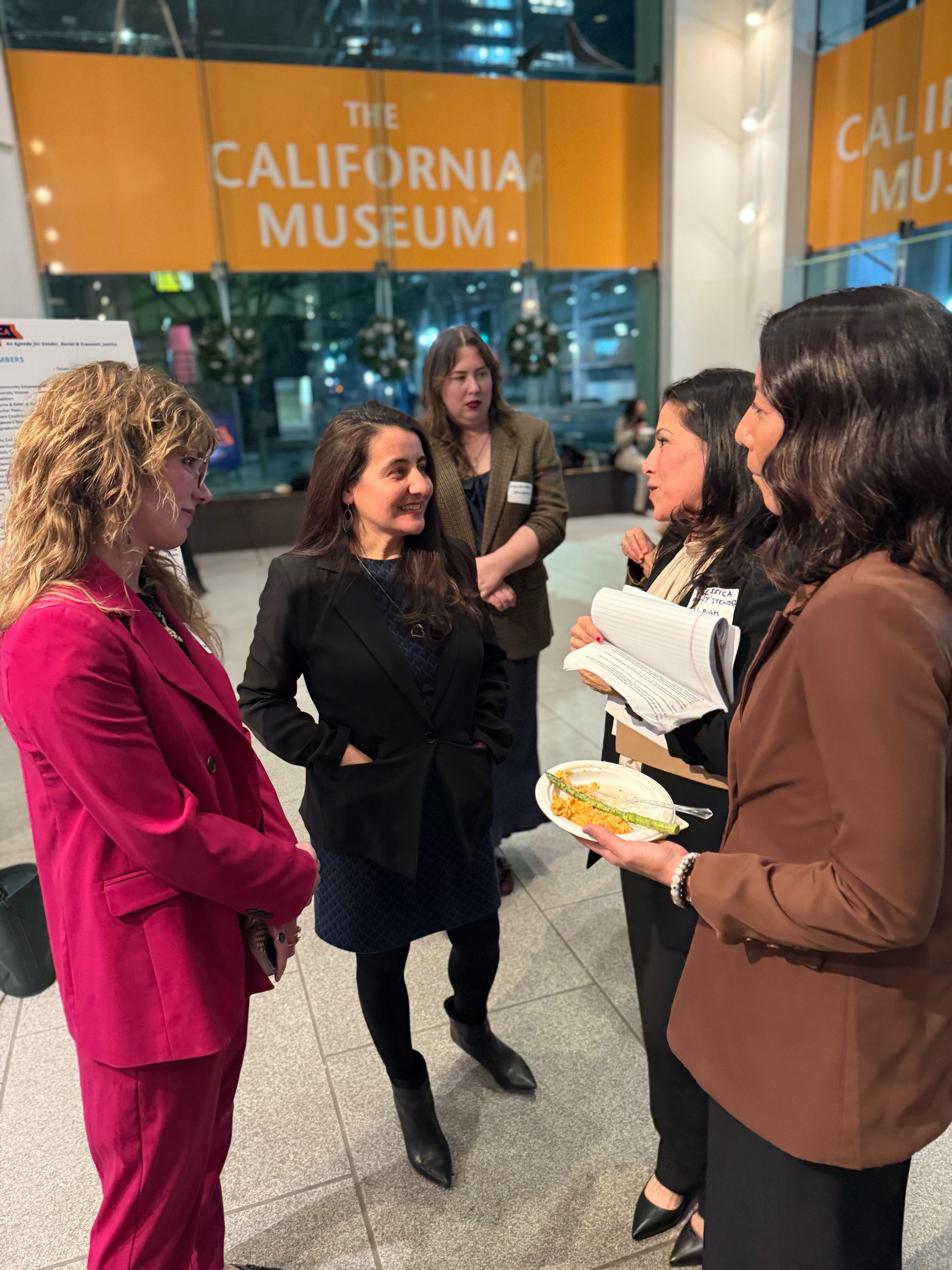 Image of women talking at the event, including a legislator from the Women's Caucus. The California Museum banner is in the background